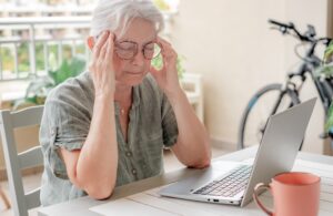 Older woman with glasses touching her temples while working at a laptop, symbolizing menopause-related brain fog; article on mitochondria and CoQ10 for brain energy.