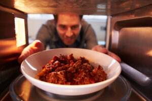 A man placing a white bowl of Paleo chili into a microwave, with a hearty beef and tomato mixture inside the bowl.