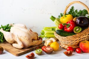 Raw whole chicken with a basket of fresh vegetables including zucchini, bell peppers, and eggplant.