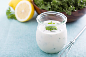 A glass jar of creamy Paleo ranch dressing garnished with fresh parsley, with lemon and greens in the background.