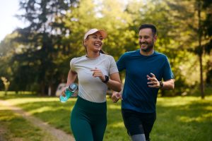 Man and woman jogging outdoors together, smiling and exercising in a park, symbolizing natural stress relief through movement.