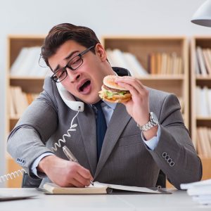 Stressed office worker multitasking while eating a fast-food burger, symbolizing stress-related comfort food cravings.